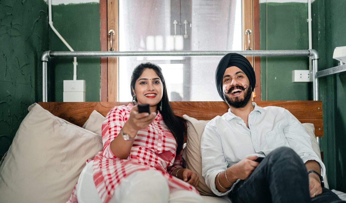 happy young indian couple watching tv in bedroom together