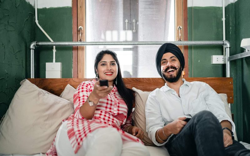 happy young indian couple watching tv in bedroom together