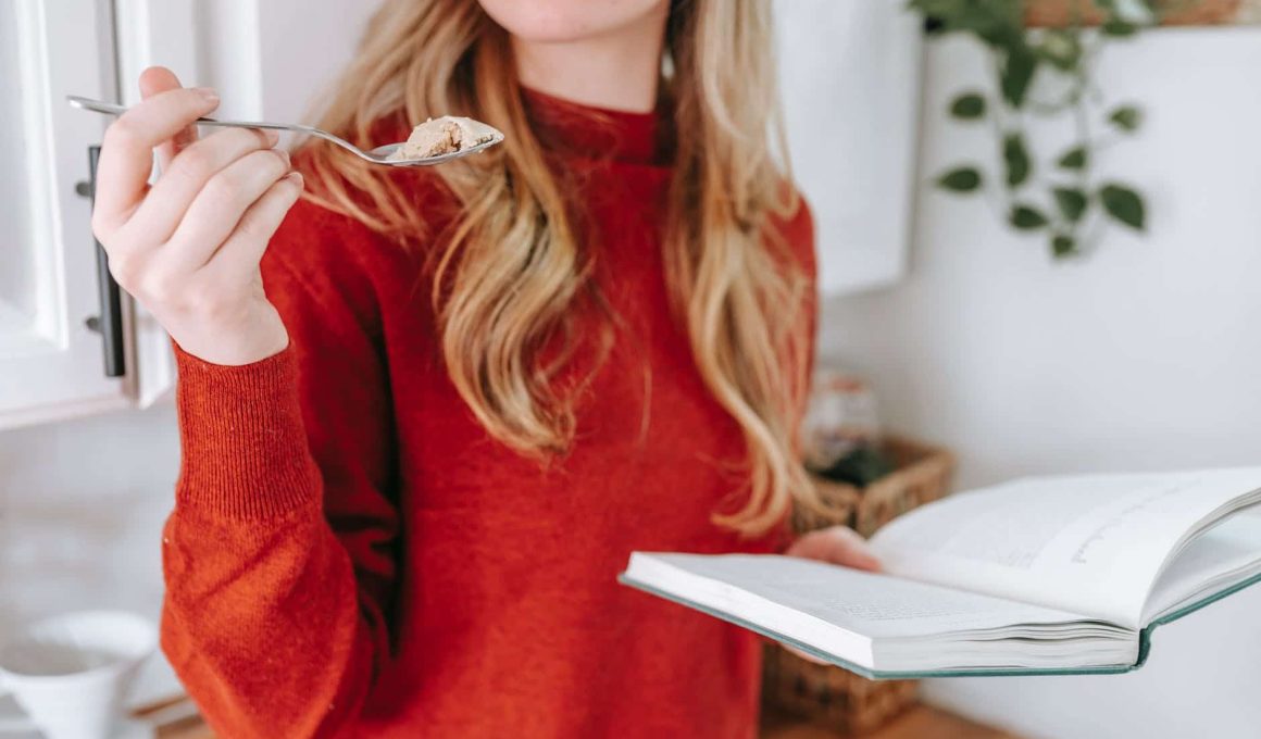 crop female student reading book and eating yogurt at home