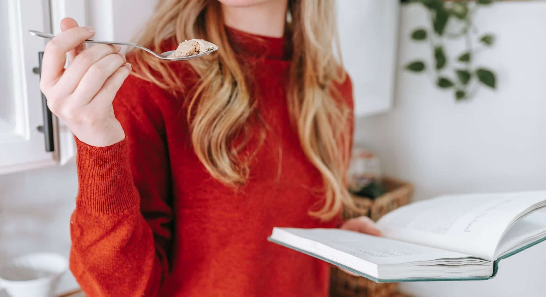 crop female student reading book and eating yogurt at home