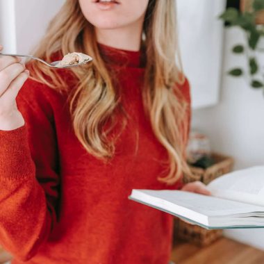 crop female student reading book and eating yogurt at home