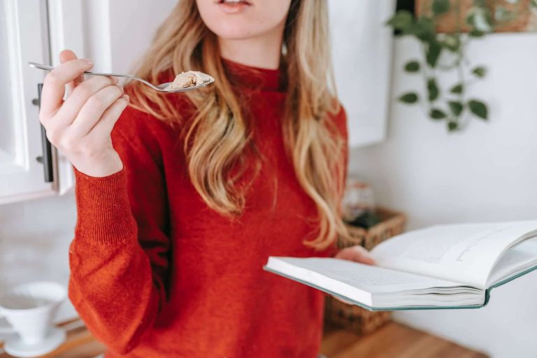 crop female student reading book and eating yogurt at home