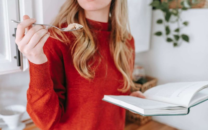 crop female student reading book and eating yogurt at home