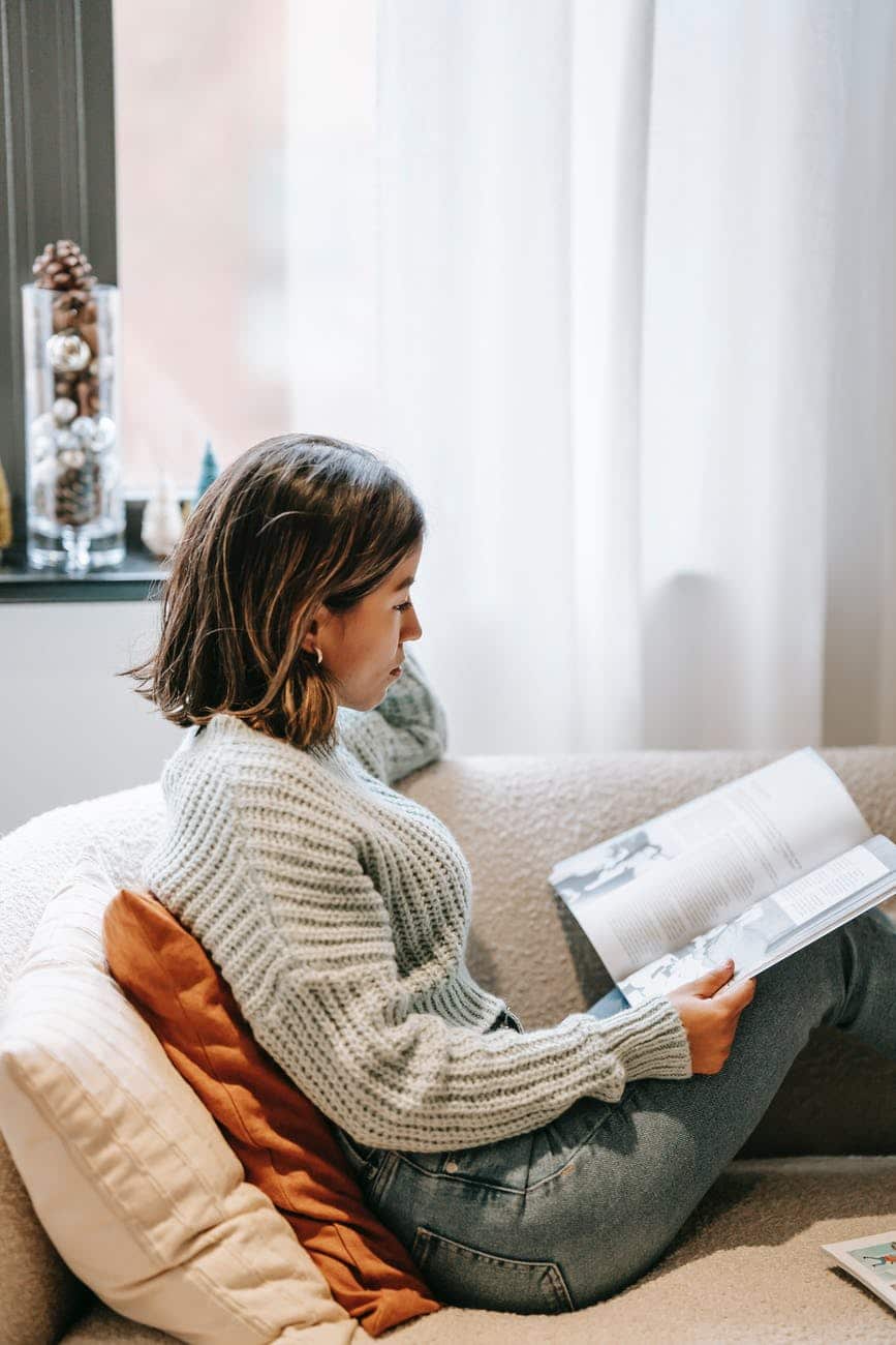 focused ethnic woman leaning on cushions and reading book