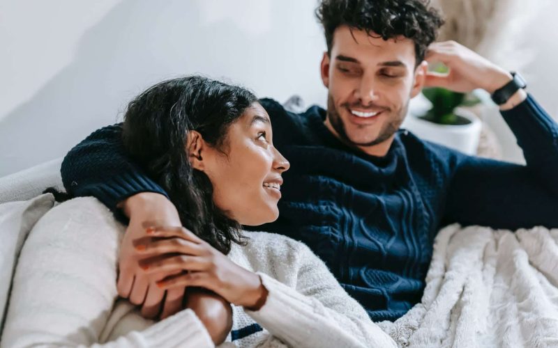 cheerful multiethnic couple resting on couch under blanket